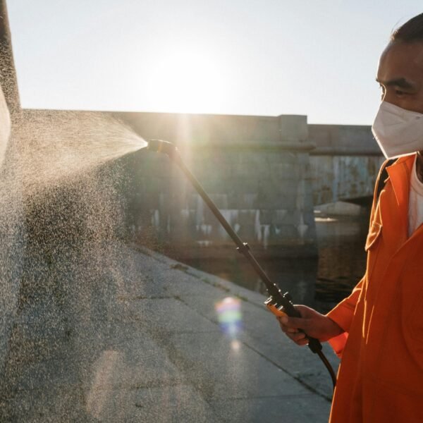 Man in orange uniform wearing facemask sprays water on wall outdoors, sunlight shining.